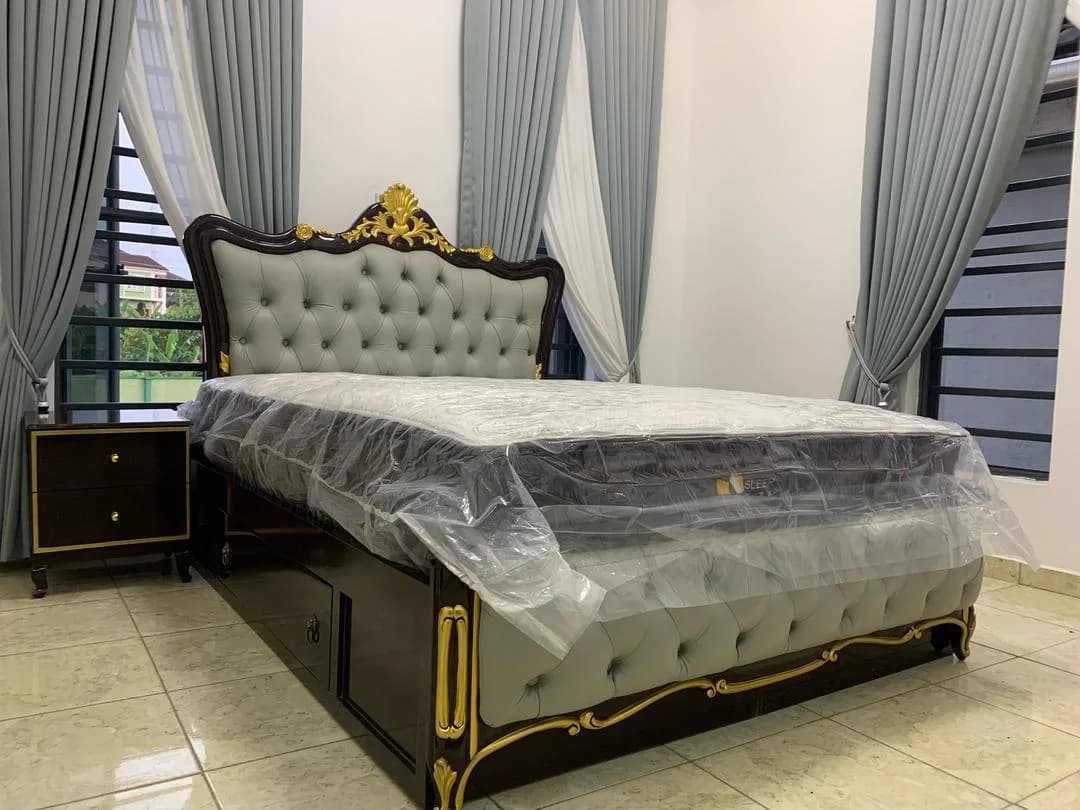 Grey tufted bed with gilded carved headboard, mattress still wrapped in plastic.