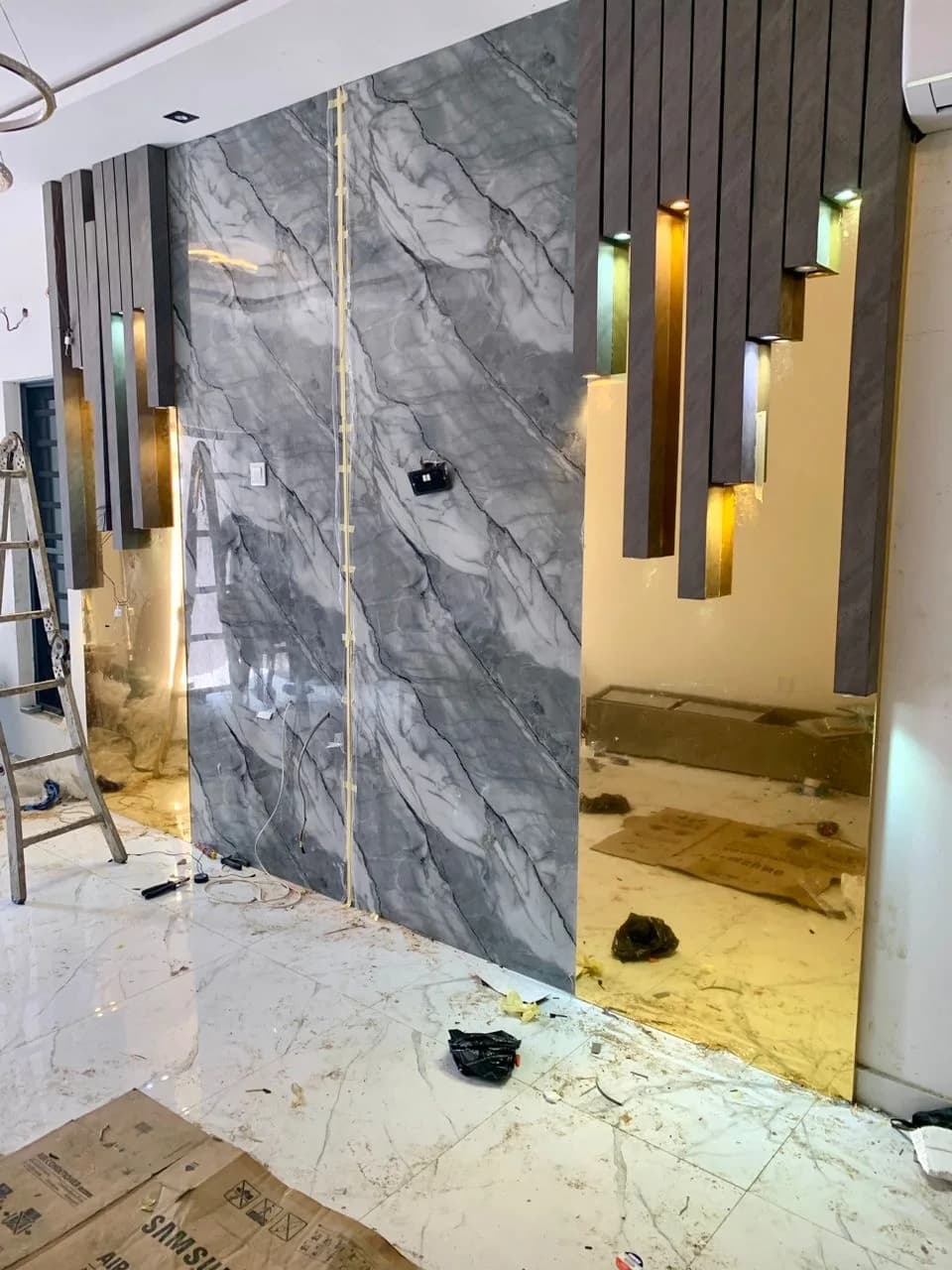 Marble slab feature wall part-clad, brass fins and timber slats going up beside a stepladder.