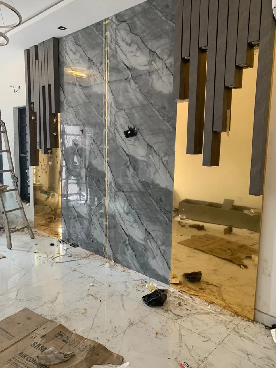Marble slab feature wall mid-installation with timber slats, gold mirror panel, ladder and floor debris.