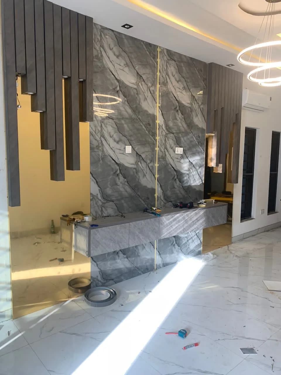 Wide view of a floating grey console and marble feature wall mid-install, with timber slats and gold mirrors.