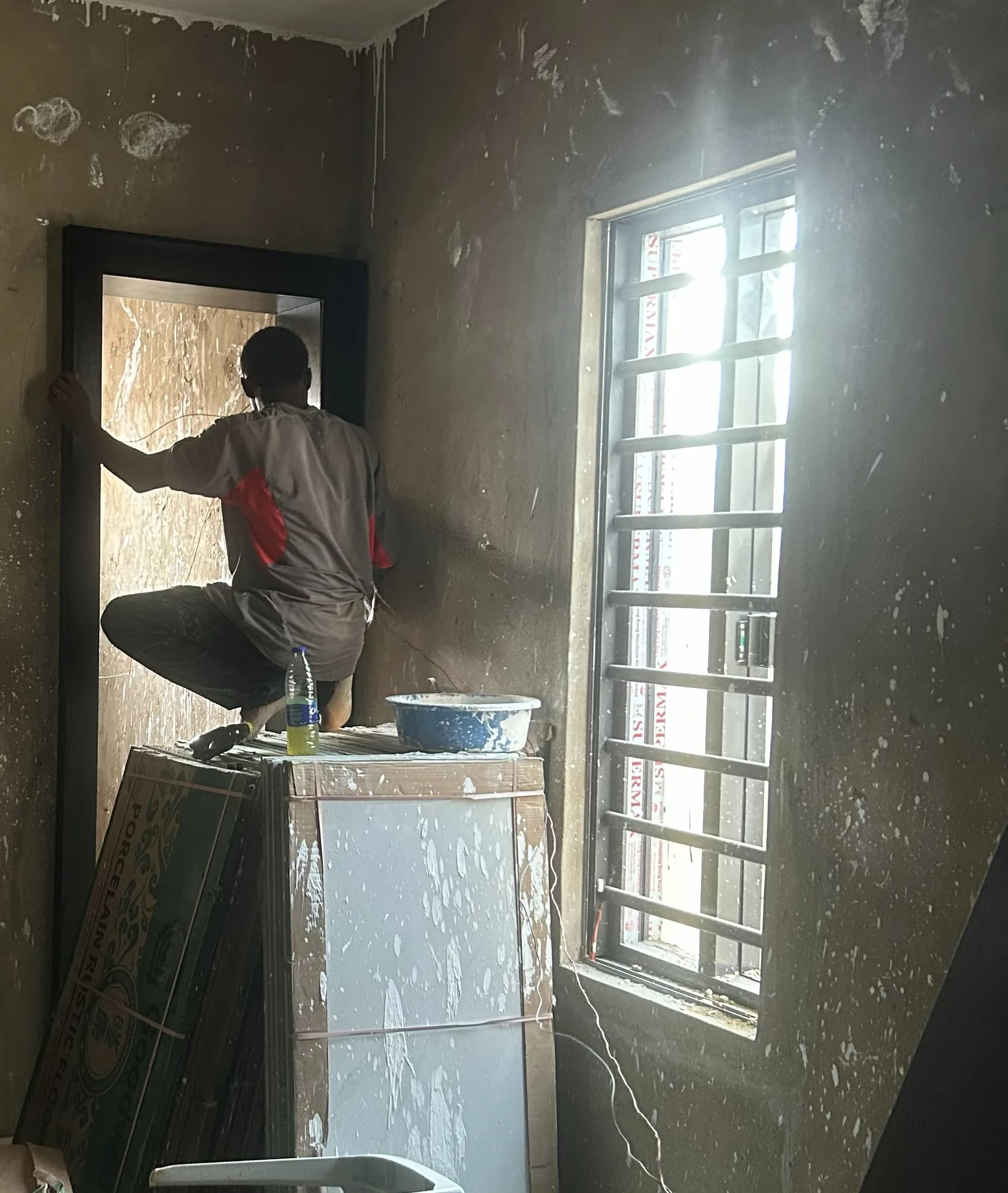 A fitter crouched on a stacked crate, setting a black door frame into a raw plastered room beside a barred window.
