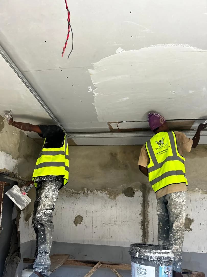 Two WoodAccent fitters in hi-vis vests skimming a ceiling on site, a plaster tub at their feet.