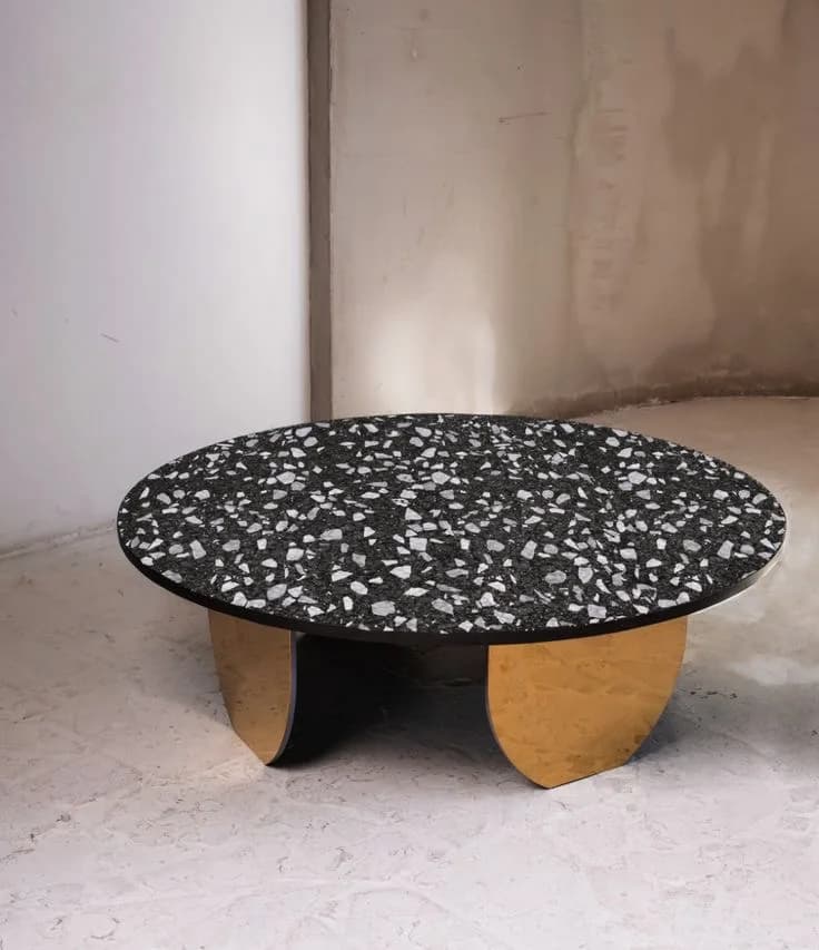 Round black terrazzo coffee table on curved brass base panels in a bare concrete room