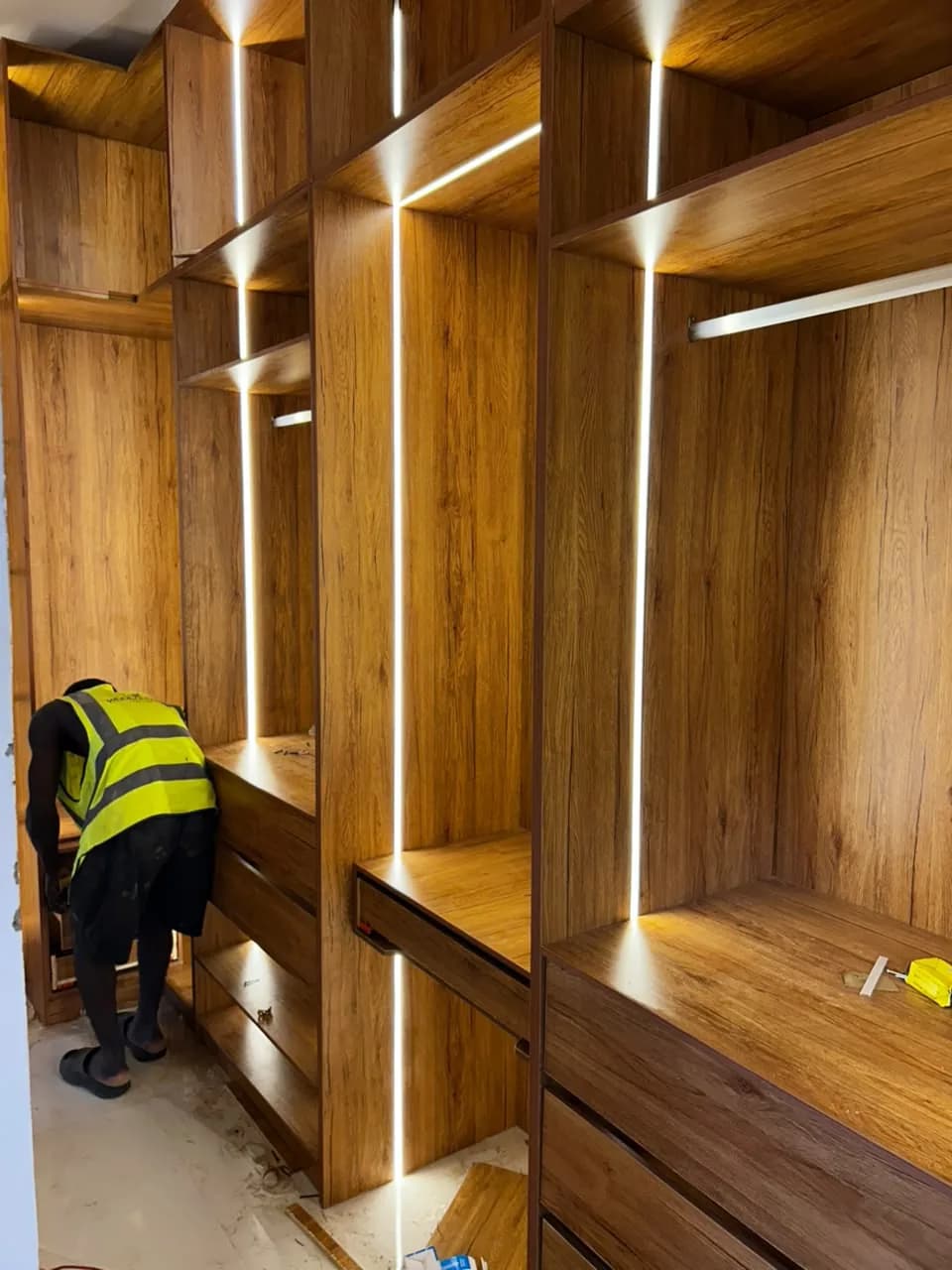 A fitter installing drawers in timber-grain wardrobe cabinetry with illuminated open compartments.