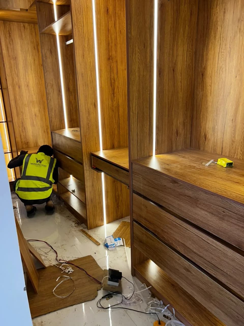 A fitter in a WoodAccent hi-vis vest crouched at the drawers of a lit timber-grain wardrobe, cables and offcuts on the floor.