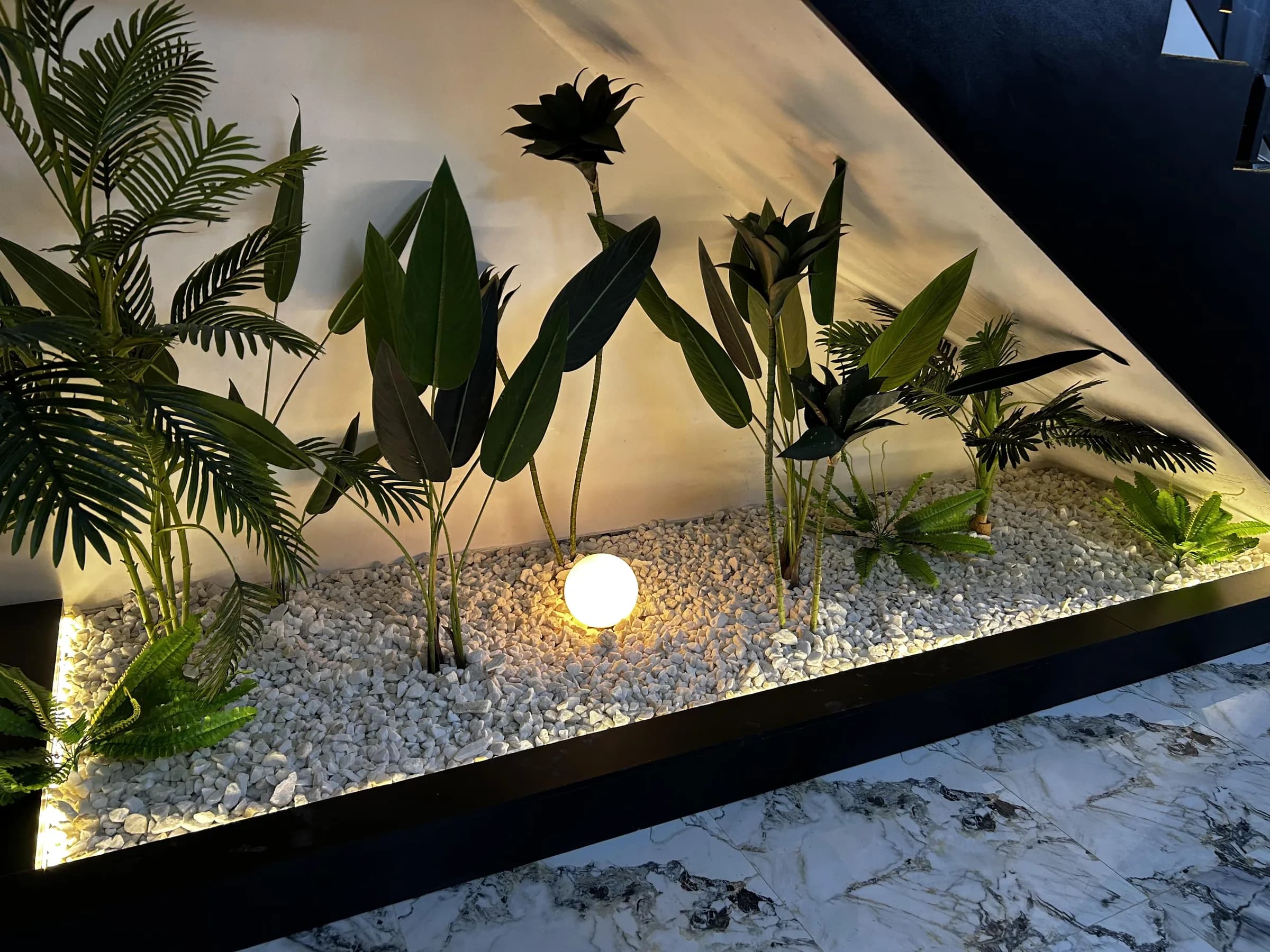 A lit planter built under the stair of a finished Lagos home, black-edged and filled with white gravel and palms.