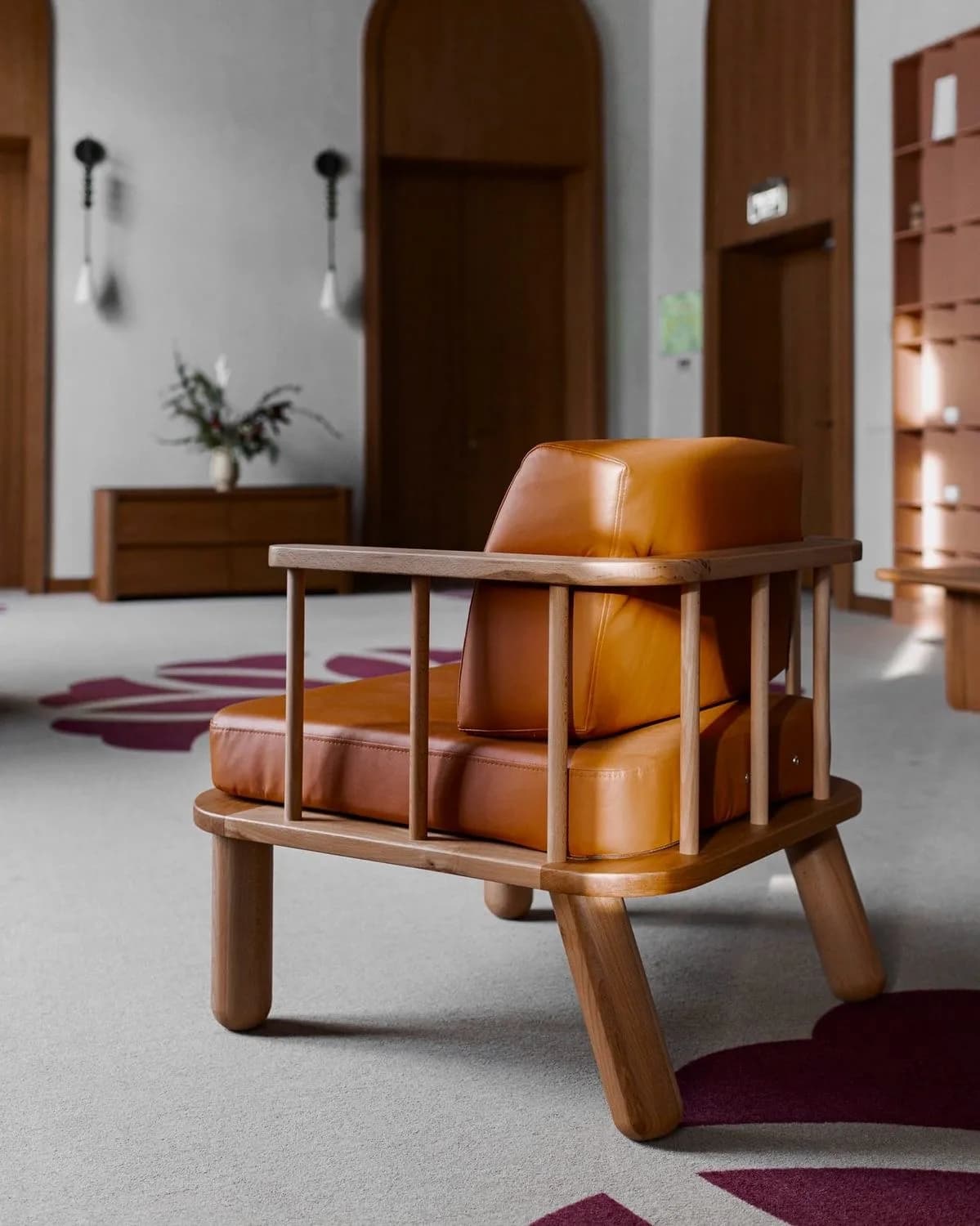 Cognac leather armchair with a pale wood spindle frame and chunky round legs on grey carpet.